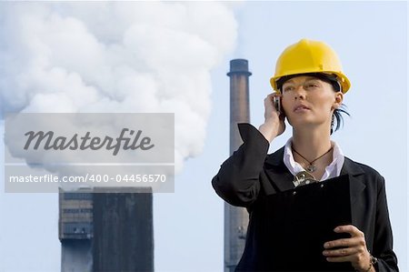 A beautiful dark haired woman wearing a hard hat and talking on her mobile phone while standing in front of a factory pumping out pollution from its chimneys