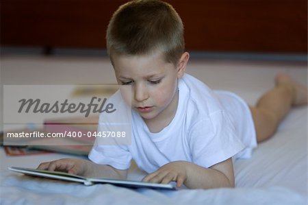 Little blond boy lying down on a bed with a book open in his hands