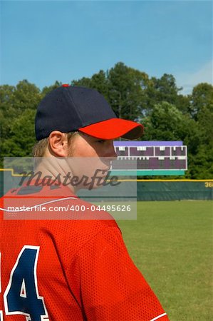 young teen male stands on baseball field in red navy uniform.  He is looking sideways.  Scoreboard is in background of picture.