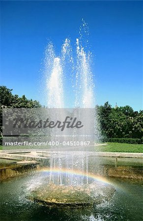 A rainbow is in the fountain of city park, powerful streams of water on a background juicy greenery and bright clean dark blue sky. Warm sun summer day