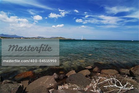 Beautiful sea scenery on Sardinia - stones in the front, sail and blue sky with clouds