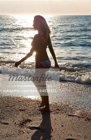 A beautiful young blond woman wearing a bikini walking along a beach at sunset