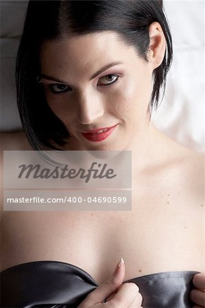Sexy naked young caucasian adult woman with red lips, short black hair and a pierced eyebrow, covered in a dark satin sheet and sitting on a bed