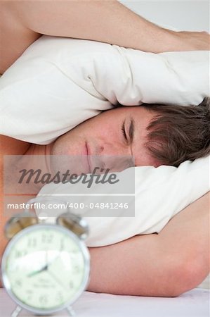 young male adult stressed by his alarm clock with his head under the pillow in the bedroom