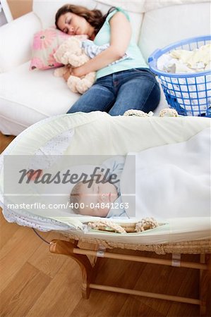 Young mother sleeping on the sofa while her baby is sleeping in his cradle in the living-room at home