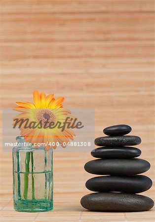 Orange snflower in a glass flask beside a black stones stack against a bamboo background