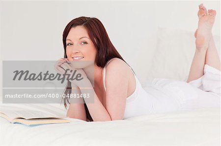 Attractive red-haired woman reading a book while lying on her bed