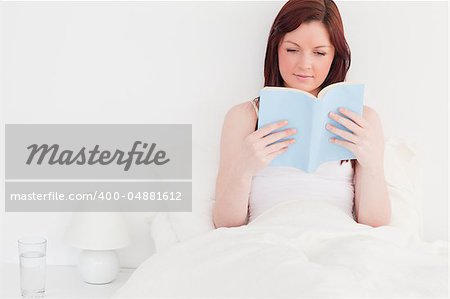 Attractive red-haired woman reading a book while sitting on her bed