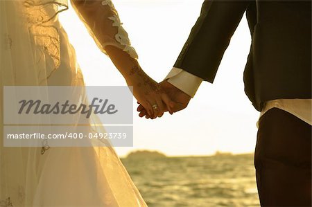 Young adult male groom and female bride holding hands on beach at sunset.