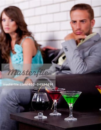 Annoyed young couple in the backround in bar or night club with colorful drinks in focus.