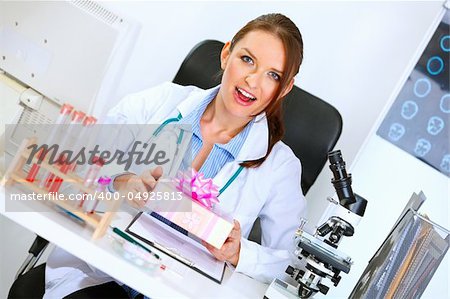 Surprised medical doctor woman sitting at office table and holding gift in hands