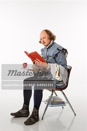 William Shakespeare in period clothing sitting in school desk reading a book.