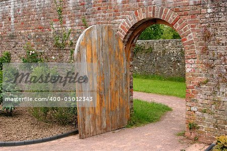 Old open arched wooden door set into an old red brick wall and leading to a grassed area beyond.