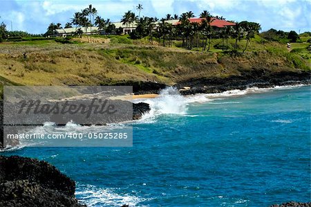 waves splash against rocks on beach below Mariott Kauai Lagoons Golf Course.  Club house sits on top of bluff.  Blue waters and green hill and black rocks.