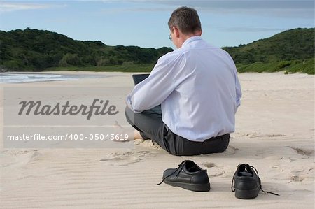Businessman working with his laptop while sitting barefoot on the beach.