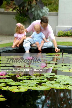 Dad sits at lilly pond with his daughter and son.  Focus is on lily with soft focus on people.