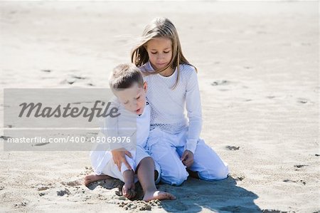 Portrait of blond sister and brother sitting in the sand dressed in white