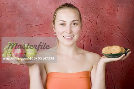Smiling young woman on a diet. Girl choosing food: fruits or cookies. Red background.