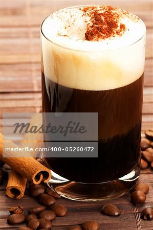 espresso in a straigt glass with milk froth cocoa powder, cinnamon sticks and coffee beans aside on wooden background