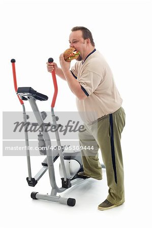 Overweight man eating a large hamburger standing by an exercising device - fittness fail concept isolated