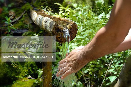 man washing his hands by spring water under wooden gutter over green natural background