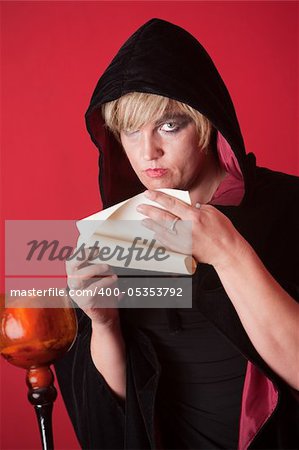 Hooded witch over red background reads a scroll
