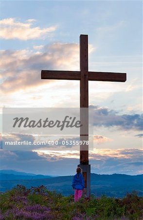 Small girl near wooden cross on summer heather flower hill top and evening country view behind