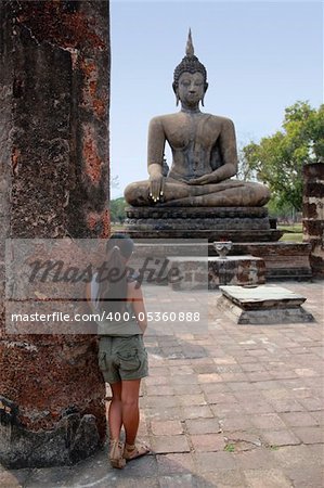 female tourist leaning against column looking up at large seated buddha statue in the temple ruins of sukhothai northern thailand