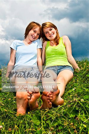 Two young teenage girl friends sitting barefoot on summer meadow