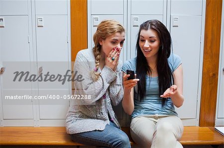 Surprised student showing a text message to her friend while sitting on a bench