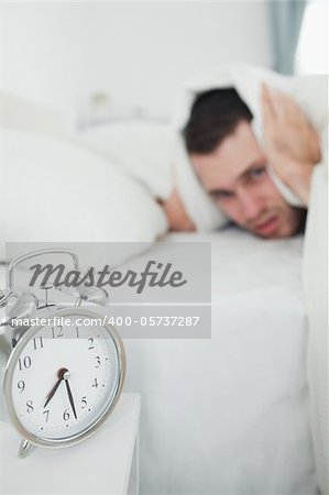 Portrait of a young man covering his ears with a duvet while his alarm clock is ringing in his bedroom