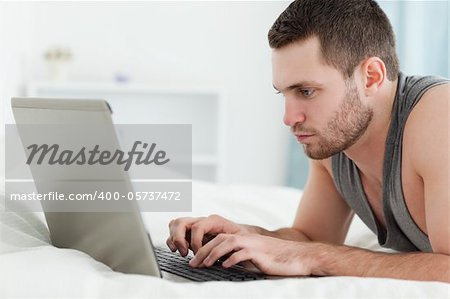 Focused man using a laptop while lying on his belly in his bedroom