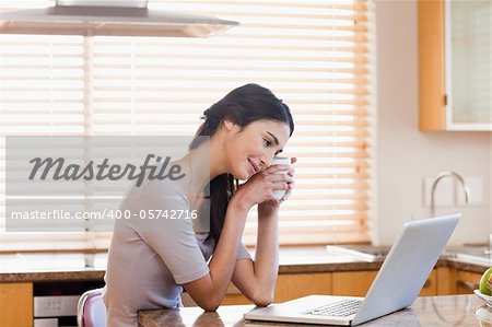 Lovely woman using a laptop while drinking a cup of a tea in her kitchen