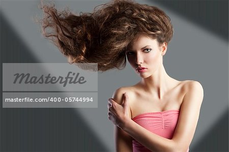 beauty fashion portrait of beautiful young brunette with curly hair flying and creative hairstyle. she looks in to the lens, her hair are raised on the right anche she takes right shoulder with left hand.