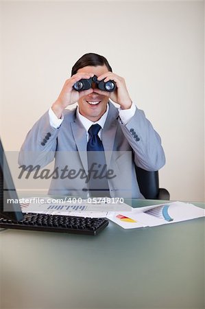 Smiling businessman at his desk looking through binoculars