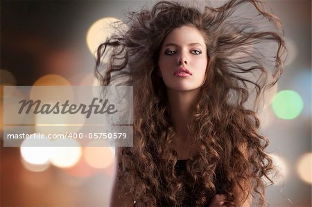 beauty fashion portrait of a very young cute alluring brunette with long curly hair with hairstyle flying in the wind and city lights