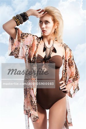 blond sexy beautiful young woman wearing a flower scarf over her body with brown swimsuit with jewellery, shw looks at right, has her right hand on the forhead and her left hand is on the left hip