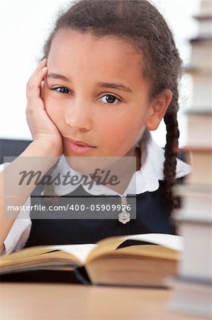 A beautiful young African American mixed race girl reading in a school classroom with a pile of books in front of her