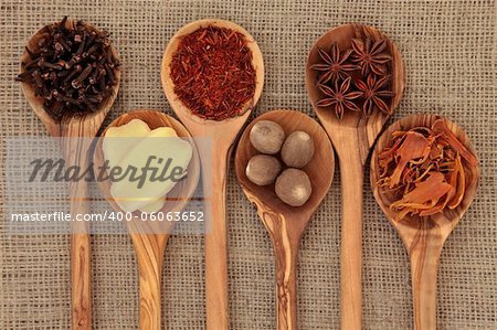 Cloves, ginger, saffron, nutmeg, star anise and mace spice in olive wood spoons over hessian background.