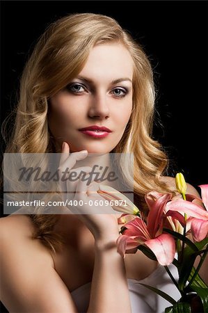 fashion portrait of a young pretty blond girl with pink lily over black background looking in camera, she is slightly turned of three quarters at left and her right hand is near the chin