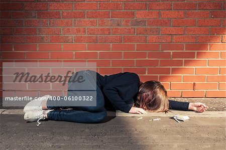 Addicted, sad young woman lying against the brick wall with syringe and cigarettes beside.  Horizontal.