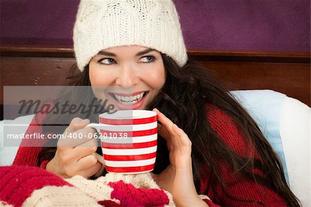 A beautiful smiling young woman enjoying a hot frink under the blankets in bed.