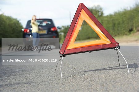 Female Driver Broken Down On Country Road With Hazard Warning Sign In Foreground