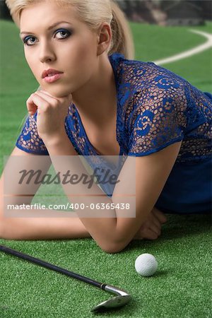 beautiful blonde girl plays golf with blue short dress and hig shoes, lying on the grass with golf-club and ball, looks in to the lens