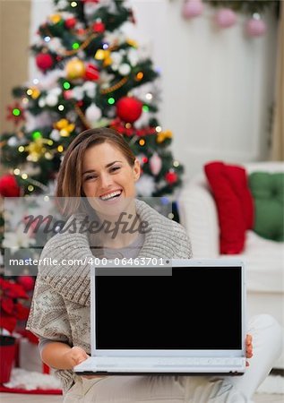 Smiling woman showing laptop blank screen near Christmas tree