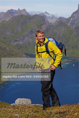 Young smiling active man with backpack hiking on Lofoten islands in Norway on sunny day high above fjord