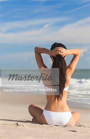 A sexy young brunette woman or girl wearing a white bikini sitting cross legged on a deserted tropical beach with a blue sky