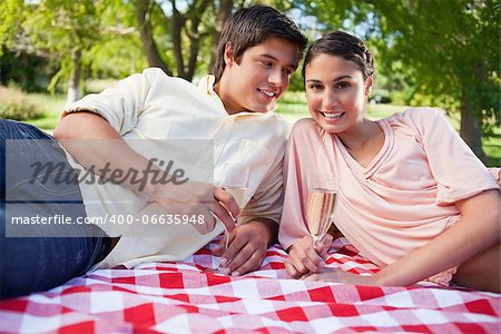 Smiling man looking at his friend while they lie down on a red and white blanket while holding glasses of champagne