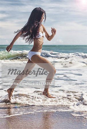 A sexy young brunette Asian woman or girl wearing a white bikini running on a deserted tropical beach with a blue sky