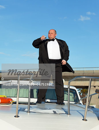 Overweight man in tuxedo standing on the deck of a luxury pleasure boat with glass red wine